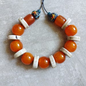 Puede incluir: Un collar hecho a mano con grandes cuentas redondas de color naranja intercaladas con cuentas rectangulares blancas. El collar también incluye cuentas más pequeñas de color naranja oscuro y cuentas decorativas con diseños azules y amarillos cerca del cierre de cordón. Sobre una superficie gris neutra.