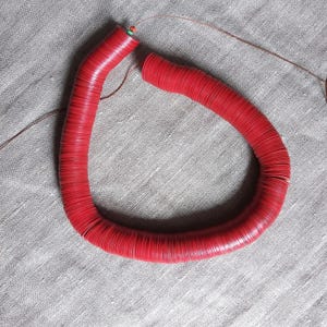 Peut inclure: Un collier rouge vif composé de nombreux petits perles circulaires et plates enfilées ensemble. Le collier est incurvé, avec une ficelle visible et un petit accent vert près du fermoir. Les perles ont un aspect légèrement texturé, et le collier repose sur un tissu de ton neutre.