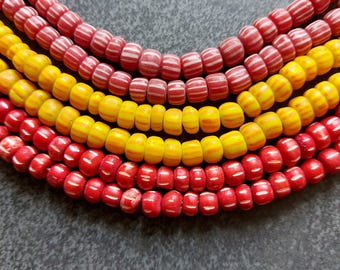 Op de afbeelding: Close-up van verschillende strengen kleurrijke kralen. De kralen zijn rood en geel, sommige met witte strepen. De kralen zijn gerangschikt in gebogen rijen, waardoor een visueel aantrekkelijk patroon ontstaat. De kralen zijn waarschijnlijk voor het maken van sieraden.