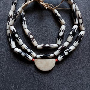 May include: A handmade necklace with white and black patterned beads. The necklace has a cream-coloured pendant and red accent beads. The beads are strung on a natural fibre cord, creating a unique and artistic accessory.