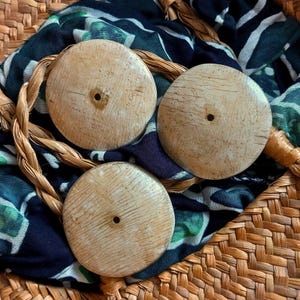 Op de afbeelding: Drie ronde, lichtbeige houten knopen met een zichtbaar nerfpatroon en een enkel gat in het midden. De knopen liggen op een donkerblauwe en groene stof met patroon, die in een gevlochten bruine rieten mand is geplaatst.