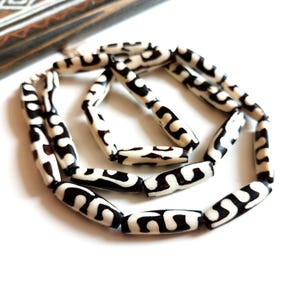 Puede incluir: Un collar con cuentas ovaladas en crema y negro. Las cuentas tienen un patrón repetitivo de remolinos y líneas negras. El collar se exhibe sobre una superficie blanca, con una parte de un objeto de madera estampada en el fondo.