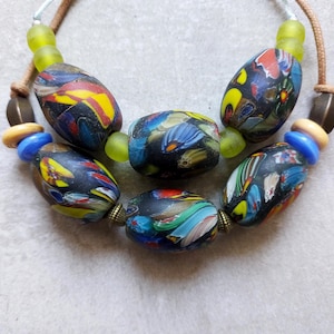 Könnte beinhalten: Eine handgefertigte Halskette mit großen, oval geformten Glasperlen mit farbenfrohen abstrakten Designs. Die Perlen sind hauptsächlich schwarz mit Akzenten in Gelb, Rot, Blau und Grün. Die Halskette enthält kleinere blaue und gelbe Perlen und ist auf einer braunen Kordel aufgefädelt.