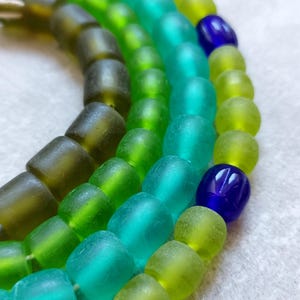 May include: Close-up of strands of colorful glass beads in shades of green and blue. The beads are oval-shaped and have a slightly irregular texture. The colors range from dark olive green to bright turquoise and deep indigo.