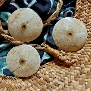 Puede incluir: Tres botones redondos de color blanquecino con un agujero central y veta visible. Los botones descansan sobre una cesta de paja tejida, con tela estampada y detalles de rafia trenzada. Los botones tienen un diámetro de aproximadamente 3,8 cm.