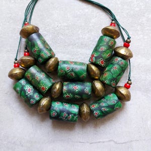 Peut inclure: Un collier fait à la main avec des perles de verre vertes et noires ornées de motifs floraux rouges. Le collier comprend également des perles en métal couleur laiton et des perles rouges, montées sur un cordon vert.