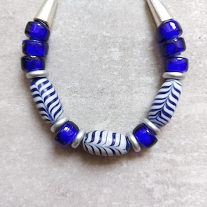 Può includere: Una collana con perline blu e bianche. La collana ha perline cilindriche blu scuro separate da anelli argentati. Le perline centrali sono bianche con un motivo blu a forma di foglia. La collana ha terminali argentati a forma di cono.