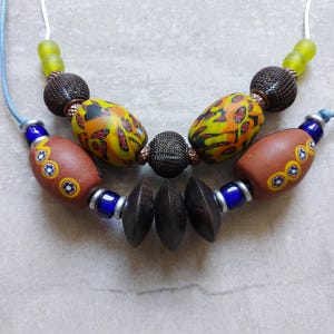 Peut inclure: Un collier artisanal composé de diverses perles. Il comprend des perles de verre marron, jaune et orange à motifs, des perles marron foncé et des perles bleues et vertes. Le collier est monté sur un cordon bleu clair.