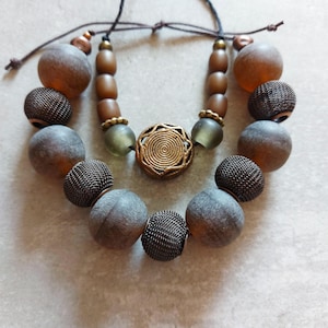 May include: A statement necklace with a mix of beads in various shapes and textures. The necklace features large, round, frosted beads in shades of brown and grey, interspersed with mesh and cylindrical beads. A central, circular, gold-toned pendant adds a decorative element.