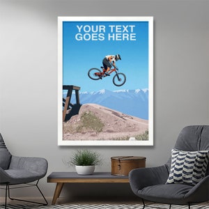 May include: A digital illustration of a mountain biker in mid-air, performing a trick on a red mountain bike. The biker is wearing a helmet and a black and white jersey. The background features a blue sky, mountains, and a wooden ramp. The text "YOUR TEXT GOES HERE" is displayed at the top of the image.