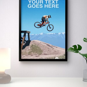 May include: A black framed poster with a blue sky and mountains in the background. A person is riding a red mountain bike over a dirt jump. The text "YOUR TEXT GOES HERE" is at the top of the poster.