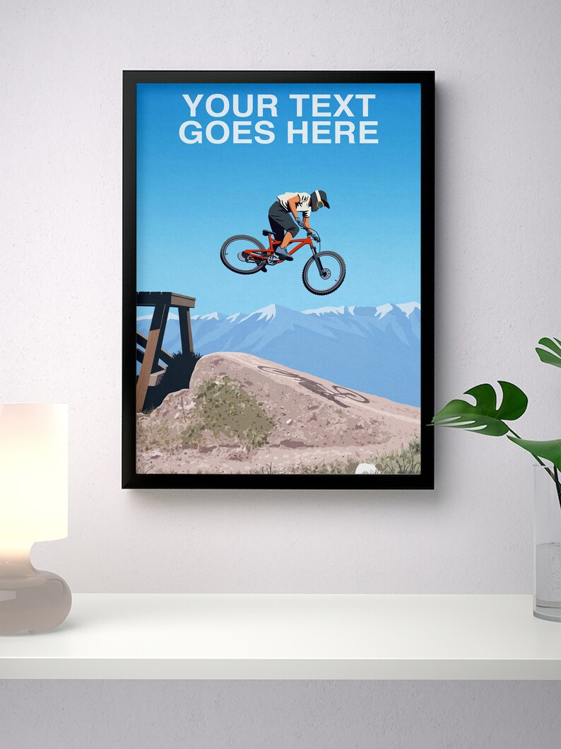 May include: A black framed poster with a blue sky and mountains in the background. A person is riding a red mountain bike over a dirt jump. The text "YOUR TEXT GOES HERE" is at the top of the poster.