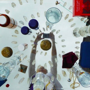 May include: A table top with a variety of crystals, glass containers, and other objects. The table is covered in a white cloth. There is a person's reflection in the table, holding a mobile phone. The text "Portals of Light" is visible on a red book.