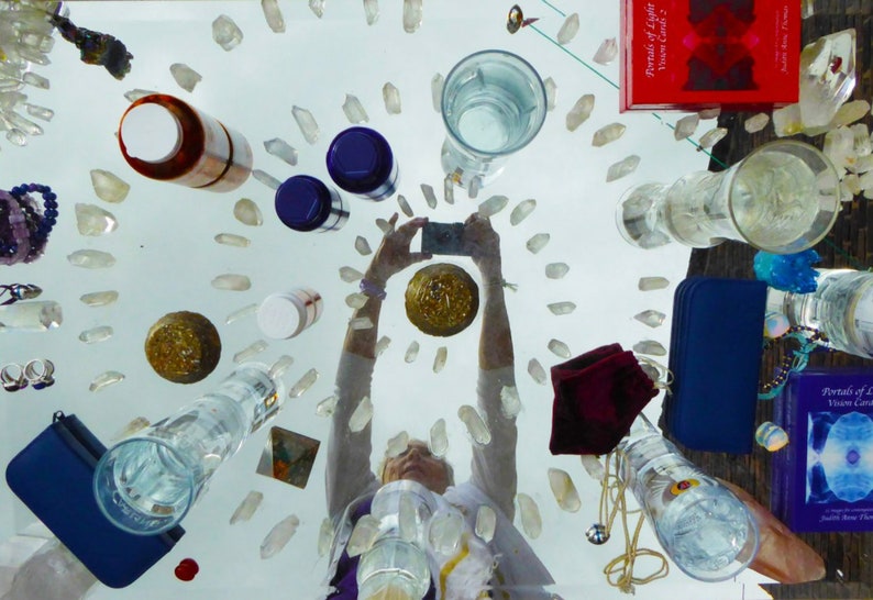May include: A table top with a variety of crystals, glass containers, and other objects. The table is covered in a white cloth. There is a person's reflection in the table, holding a mobile phone. The text "Portals of Light" is visible on a red book.