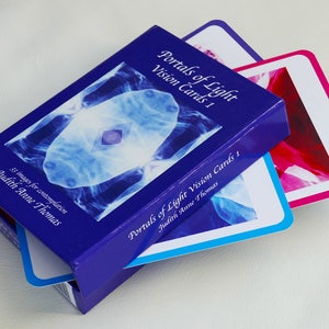 May include: A blue box with the title "Portals of Light Vision Cards 1" by Judith Anne Thomas. The box contains 33 images for contemplation. Two cards are partially visible, one with a blue and white image and the other with a pink and white image.