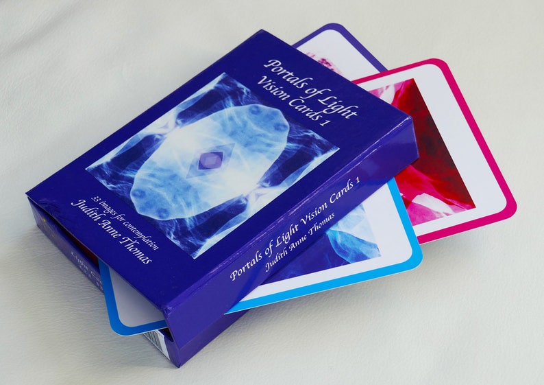 May include: A blue box with the title "Portals of Light Vision Cards 1" by Judith Anne Thomas. The box contains 33 images for contemplation. Two cards are partially visible, one with a blue and white image and the other with a pink and white image.