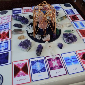 May include: A white table with a gold geometric structure in the centre.  Surrounding the structure are various crystals and cards with images and words like "bliss", "grace", "truth", and "inspiration".