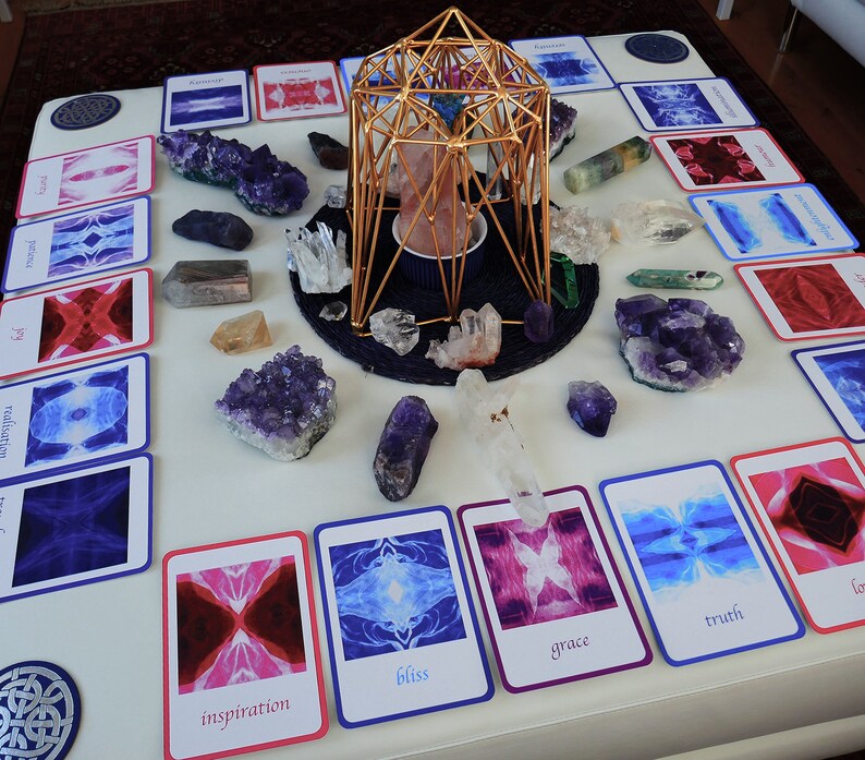 May include: A white table with a gold geometric structure in the centre.  Surrounding the structure are various crystals and cards with images and words like "bliss", "grace", "truth", and "inspiration".