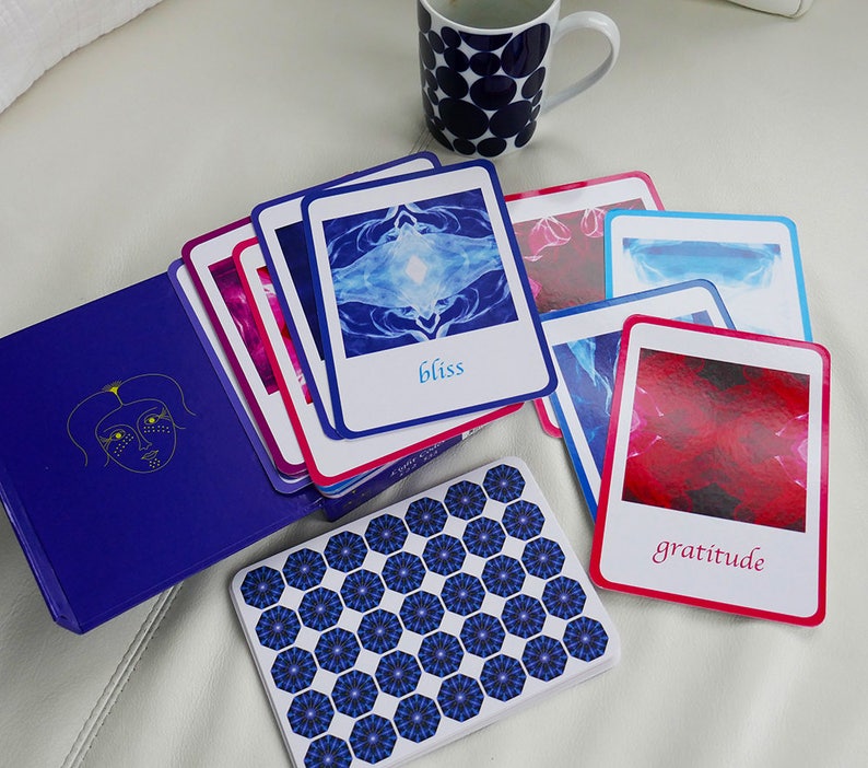 May include: A set of affirmation cards with colourful abstract designs and inspirational words such as "bliss" and "gratitude". The cards are arranged on a white surface with a blue and white patterned mug in the background.