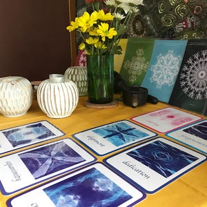 May include: A table with a yellow tablecloth and a variety of cards with blue and white designs. The cards have words like "equanimity", "dedication", and "alignment" printed on them. There are also two white ceramic candle holders and a vase with yellow flowers.