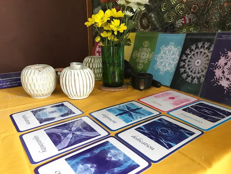 May include: A table with a yellow tablecloth and a variety of cards with blue and white designs. The cards have words like "equanimity", "dedication", and "alignment" printed on them. There are also two white ceramic candle holders and a vase with yellow flowers.