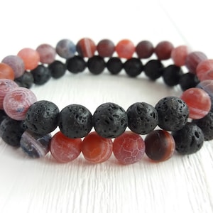 May include: Two beaded bracelets. One bracelet features black lava beads, and the other features red, pink, and purple agate beads. The bracelets are stacked, showcasing the contrast in textures and colours.