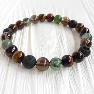 Könnte beinhalten: Ein Armband mit verschiedenen Natursteinperlen in Braun-, Grün- und Schwarztönen. Das Armband enthält Tigerauge, Lava und andere Edelsteinperlen, die auf einer elastischen Schnur aufgefädelt sind. Das Armband wird auf einer weißen Oberfläche präsentiert.