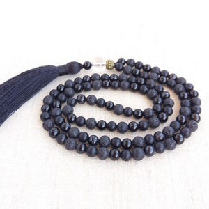 May include: A mala necklace featuring alternating black lava stone beads and smooth black onyx beads. The necklace includes a clear crystal bead, a small metallic spacer, and a long black tassel attached at the top of the circular strand.