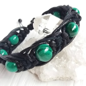 Könnte beinhalten: Ein schwarzes geflochtenes Armband mit grünen Malachitperlen. Das Armband ist mit einem Knoten gebunden und liegt auf einer weißen Oberfläche.