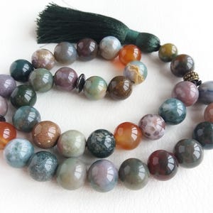 May include: A necklace made of colourful, round gemstones in shades of green, brown, orange, and purple. The necklace has a dark green tassel attached to it.