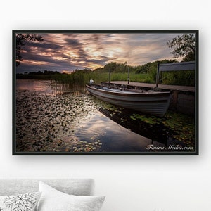 May include: A wooden rowboat is docked at a wooden pier on a lake. The lake is covered in lily pads and the sky is a vibrant orange and pink sunset.