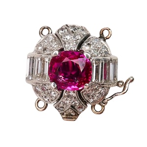 May include: A detailed view of a jewellery clasp with a central, large, round, deep pink gemstone. The clasp is encircled by smaller, clear gemstones and rectangular-cut stones, set in a silver-coloured metal. It features a hook closure.