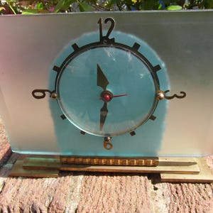 May include: A vintage Art Deco clock with a rectangular frosted glass face and a light blue clock face. The clock has a brass-coloured frame and base, with black clock hands and a red second hand. The clock is approximately 20 cm wide.