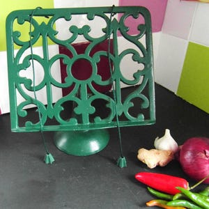 May include: A dark green cast iron cookbook stand featuring an intricate openwork scroll pattern. The stand includes a circular base and two thin green cords with weighted ends hanging from the top frame to hold pages open while cooking.