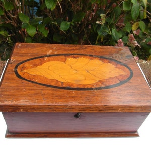 May include: A wooden box with a dark brown finish and a marquetry inlay of a leaf in an oval shape. The box has a hinged lid and a metal latch.