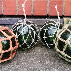 May include: Four green glass fishing floats with netting. The floats are various colours, including orange, white, and natural rope. Each float has a loop for hanging. The background includes a brick wall and concrete surface.