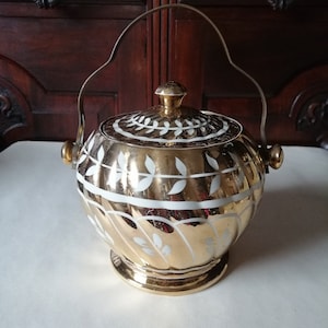 May include: A gold-coloured, lidded serving dish with a handle. The dish has white leaf patterns and a matching gold knob on the lid. The handle is metal. The dish is likely vintage and suitable for serving food.