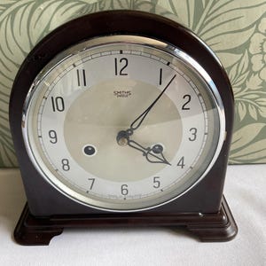 May include: A dark brown mantel clock with a rounded top and a chrome bezel. The clock face is cream-coloured with black Arabic numerals and the text Smiths Enfield. It features two black hands and two winding holes on the lower half.