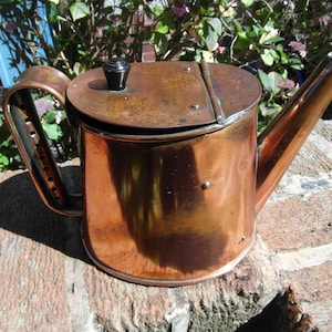 May include: An antique copper teapot with a hinged lid and a long spout. The teapot features a curved handle and a black knob on the lid. The copper has a warm, aged patina, indicating its age. The teapot is on a brick surface.