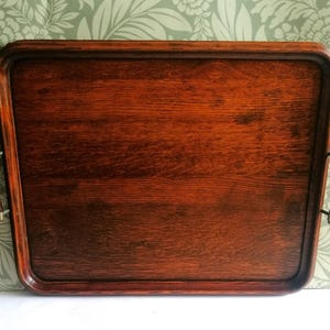 May include: A rectangular wooden serving tray with a dark brown finish and visible wood grain. The tray features rounded corners and two curved metal handles attached to the sides, each with a dark wooden grip for ease of handling and transport.