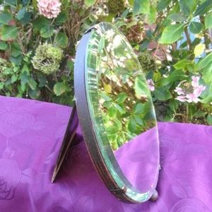 May include: A vintage oval-shaped hand mirror with a wooden frame and a bevelled glass mirror. The mirror is standing on a purple surface.