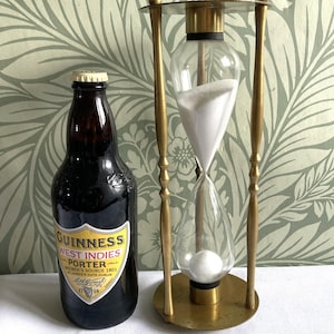 May include: A dark brown glass Guinness West Indies Porter beer bottle with a yellow and white label next to a brass hourglass filled with white sand. The bottle label features the text Guinness West Indies Porter Brewer's Source 1801.