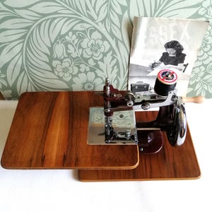 May include: A vintage sewing machine with a wooden base and a dark maroon body. A spool of black thread sits on top, next to a manual. The machine has a silver needle and a wooden handle. The base is made of two wooden platforms.