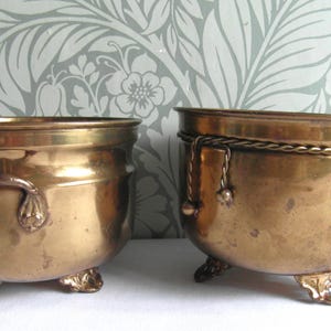 May include: Two antique brass planters. The planters have a rounded shape, decorative handles, and small feet. One planter has a rope detail around the rim. The brass has a warm, aged patina, suggesting vintage craftsmanship.