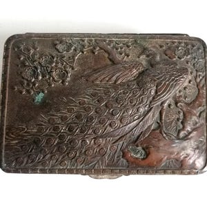 May include: A rectangular antique metal box with a detailed relief of a peacock. The box is bronze-coloured with darker accents, and the peacock's tail feathers are prominently displayed. Floral and branch details frame the bird.