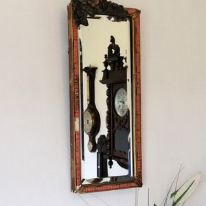 May include: A vintage mirror with a carved wooden frame and a red and gold paint finish. The mirror reflects a clock and a barometer.