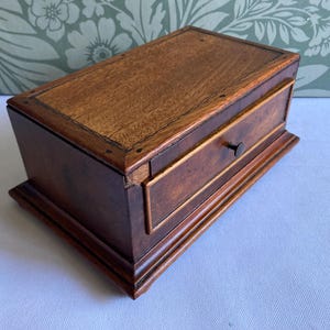 May include: A rectangular wooden box with a dark brown finish and a visible wood grain pattern. The box features a single front drawer with a small, centred black knob. The top surface is flat with a defined border, and the base has a stepped edge.