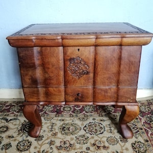 May include: A vintage wooden chest with a carved floral design and keyhole. The chest has a rectangular top with decorative edging and curved legs. The wood has a warm, brown tone. The chest is likely made of oak.