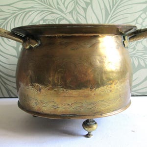 May include: A round brass pot featuring two side handles and three small feet. The exterior surface is decorated with engraved floral and wave-like patterns. The metal has a polished finish with visible signs of age and patina throughout the body.