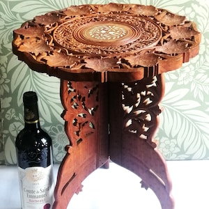 May include: A round, brown wooden folding table with intricate floral carvings on the top and legs, featuring a central inlaid floral design. Beside it stands a dark glass wine bottle with a label reading Comte de Saint Emmanuel Bordeaux.